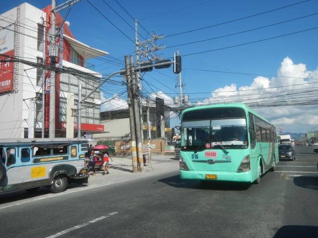 Ein grüner Bus fährt auf einer von Hochhäusern gesäumten Stadtstraße, während Passanten mit Regenschirmen auf dem Gehweg gehen und Strommasten mit Drähten am Straßenrand zu sehen sind.
