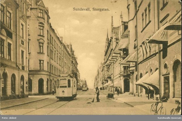 Ein altes Foto einer Stadtstraße mit einer Tram, Gebäuden mit Fenstern, Menschen, die gehen, Fahrrädern und einem bewölkten Himmel, mit Text oben und unten auf dem Bild.
