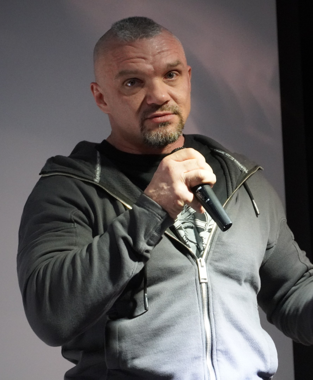 Komiker in einem schwarzen Anzug und weißem Hemd, der ein Mikrofon hält und selbstbewusst vor einer Wand steht.