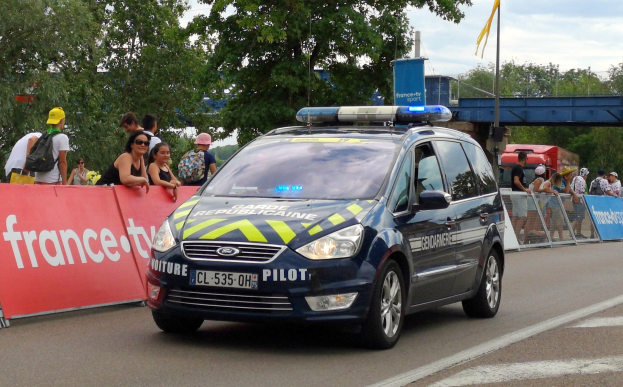 Polizeiauto fährt an einer Menge mit Schildern, Geländern, Bäumen, einer Brücke, einer Fahne und einem bewölkten Himmel vorbei.