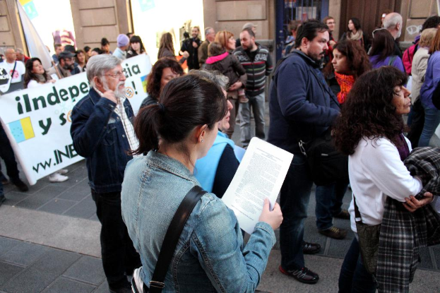 Eine Gruppe von Menschen in einem Raum, einige halten ein Banner mit Text, eine Frau trägt eine Tasche und Papiere, eine andere hält einen Mantel.