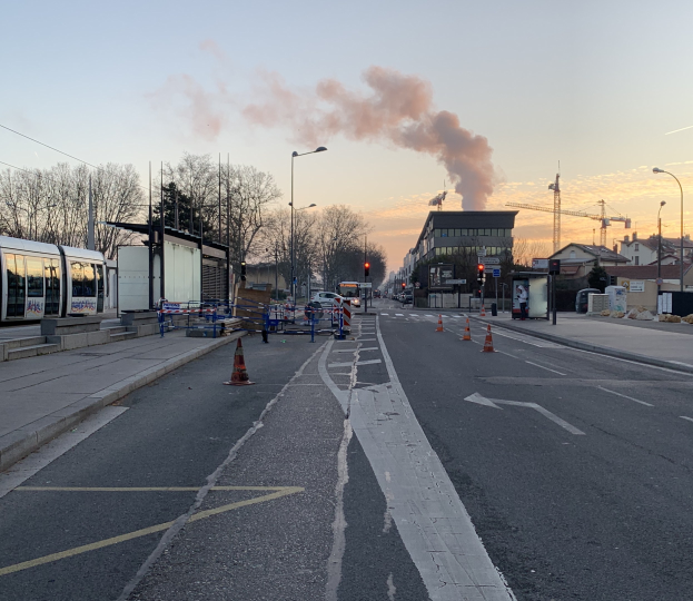 Eine Stadtstraße mit Verkehrskegeln, einem Bus, Laternen, Bäumen, Gebäuden, Kränen und Rauch, der aus einem fernen Gebäude aufsteigt.