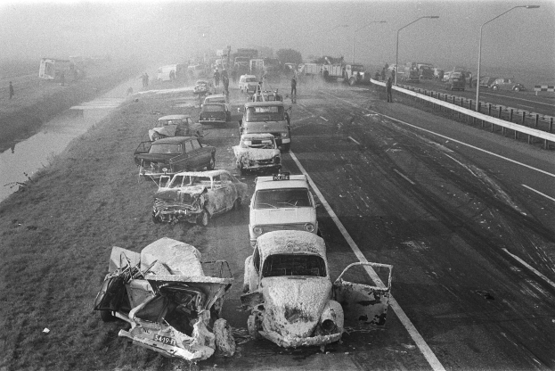Ein Schwarz-Weiß-Foto von mehreren verunglützten Autos an der Straße bei Nacht, mit Laternen, nahestehenden Menschen, Bäumen und einem Gewässer im Hintergrund.