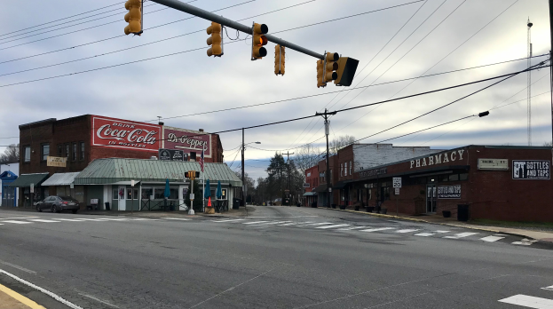 Eine Stadtstraßenkreuzung mit Ampel, Fahrzeugen, Gebäuden mit Namensschildern, Strommasten mit Kabeln, Bäumen und einem bewölkten Himmel.