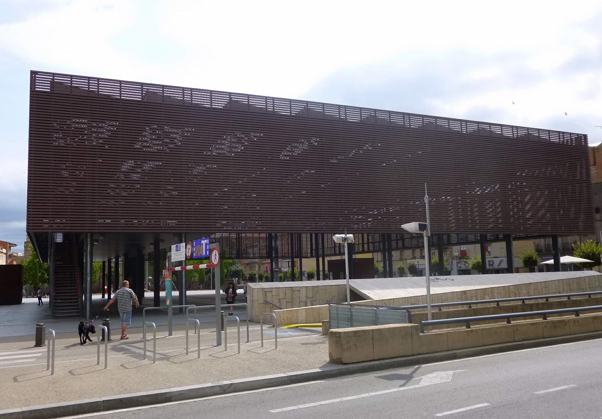 Großes modernes Gebäude mit zahlreichen Fenstern, das den neuen Eingang zur City bildet, flankiert von Straßeninfrastruktur, Fahrzeugen, Fußgängern und Grünflächen unter einem bewölkten Himmel.