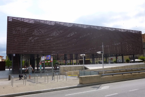 Großes modernes Gebäude mit zahlreichen Fenstern, das den neuen Eingang zur City bildet, flankiert von Straßeninfrastruktur, Fahrzeugen, Fußgängern und Grünflächen unter einem bewölkten Himmel.