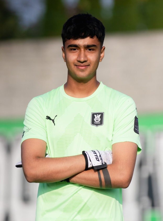 Ein junger Mann in einem grünen Trikot steht auf einem Fußballfeld mit verschränkten Armen und lächelt, mit einem unscharfen Hintergrund.