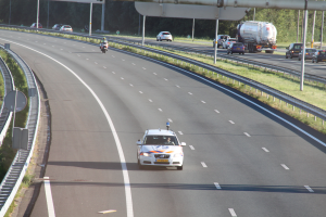 Ein Auto fährt auf einer Autobahn mit einem polizeiauto am rand, andere fahrzeuge, schienen, mäste, plätze, bürgen und bäume im hintergrund.