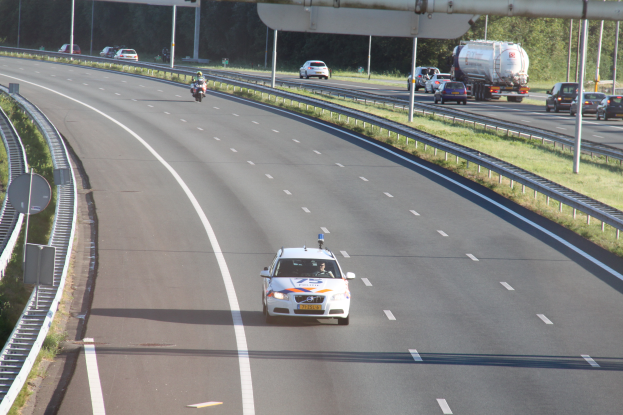 Ein Auto fährt auf einer Autobahn mit einem polizeiauto am rand, andere fahrzeuge, schienen, mäste, plätze, bürgen und bäume im hintergrund.