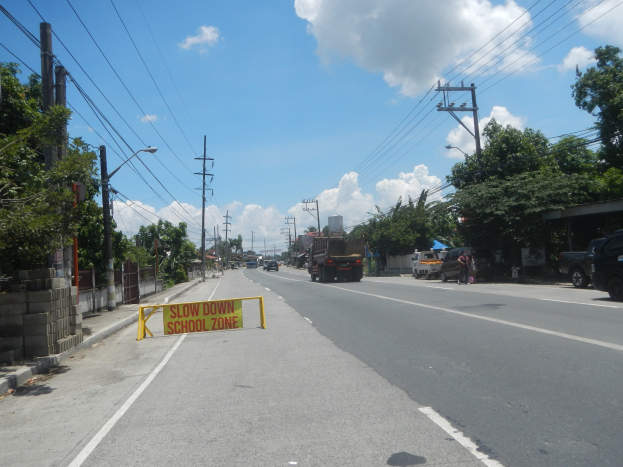 Straße mit Fahrzeugen, ein "Slow Down School Zone"-Schild, Strommäste mit Drähten, Bäume, Laternenmästele und ein bewölkter Himmel im Hintergrund.