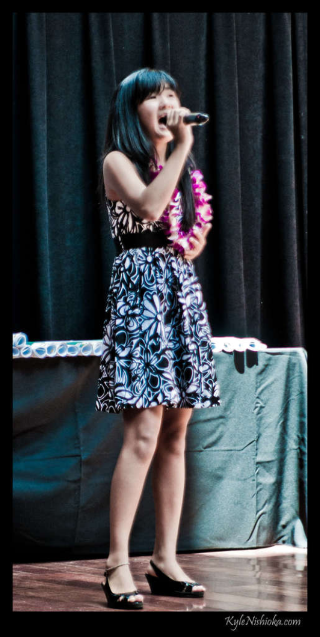 Eine Frau in einem bunten Kleid singt in ein Mikrofon auf einer Bühne mit einem Vorhang und einem Tisch mit Papieren im Hintergrund.