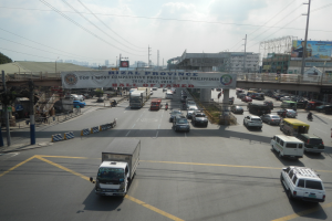 Eine belebte Straße mit zahlreichen Autos und Lastwagen, eine Brücke im Hintergrund, Strommasten mit Kabeln, ein Schild mit Text, Gebäude, Bäume und ein bewölkter Himmel.