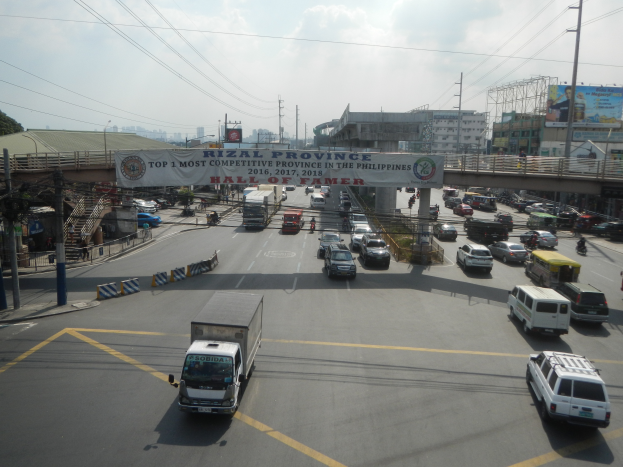 Eine belebte Straße mit zahlreichen Autos und Lastwagen, eine Brücke im Hintergrund, Strommasten mit Kabeln, ein Schild mit Text, Gebäude, Bäume und ein bewölkter Himmel.