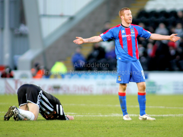 Ein Stadion mit einer Person, die steht, und einer anderen, die auf dem Rasen kniet, sowie einer Gruppe von Menschen im Hintergrund und Text auf Tafeln dahinter.