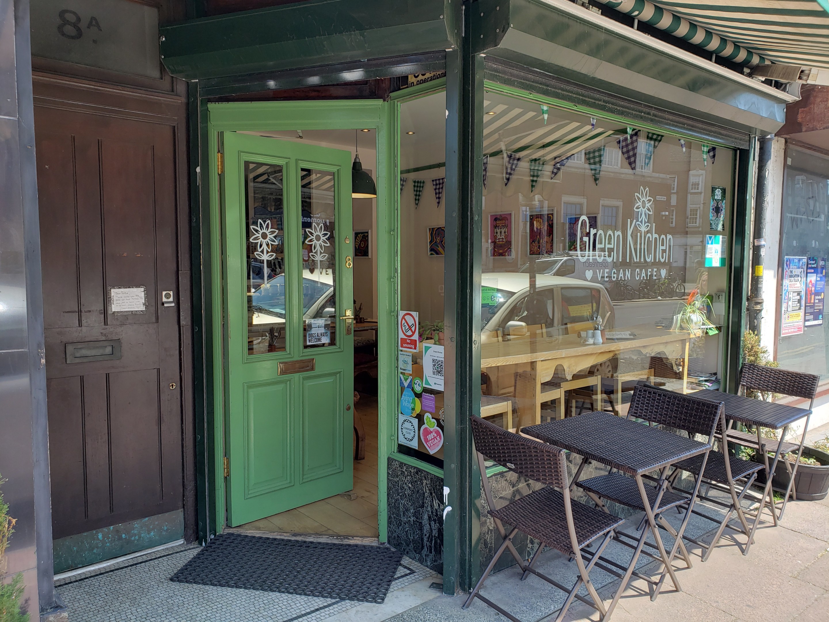 Außensitzbereich des Green Kitchen Vegan Cafe mit Tischen, Stühlen, Pflanzen, einer Fußmatte und einer Glastür mit Postern. Drinnen sind Tische, Stühle und Deckenleuchten zu sehen.