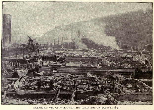 Schwarzes und weißes Foto einer Stadt nach einer Katastrophe am 5. Juni 1892, das Gebäude, Pfosten, Rauch, einen Hügel im Hintergrund und Text unten zeigt.