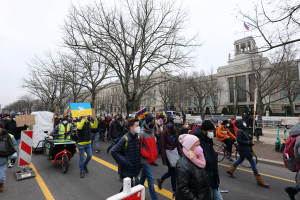 Eine große Gruppe von Menschen nimmt an einer Protestmarsch auf einer Straße in Washington, D.C., teil, einige halten Plakate und Banner, andere fahren Fahrräder, und es gibt Schilder, Bäume und einen klaren blauen Himmel im Hintergrund.