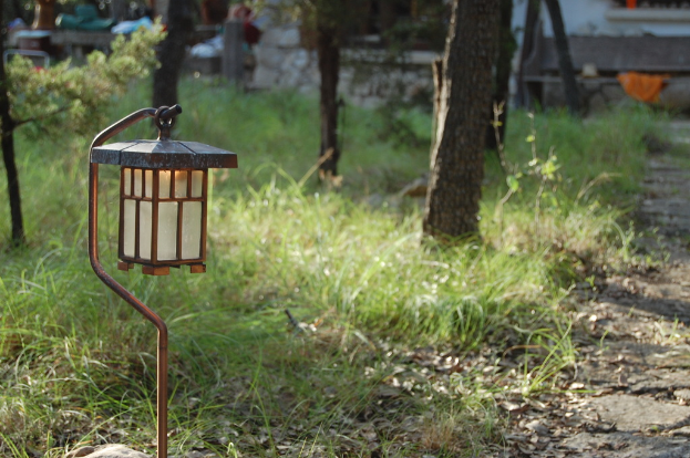 Ein Licht ist an einer Eisenstange auf der linken Seite befestigt, mit Gras unten und Pflanzen und Gebäuden oben.