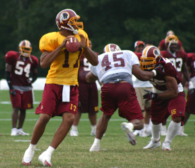Eine Gruppe von Fußballspielern in Trikots, Helmen und weißen Schuhen auf einem grünen Rasen mit Bäumen im Hintergrund.