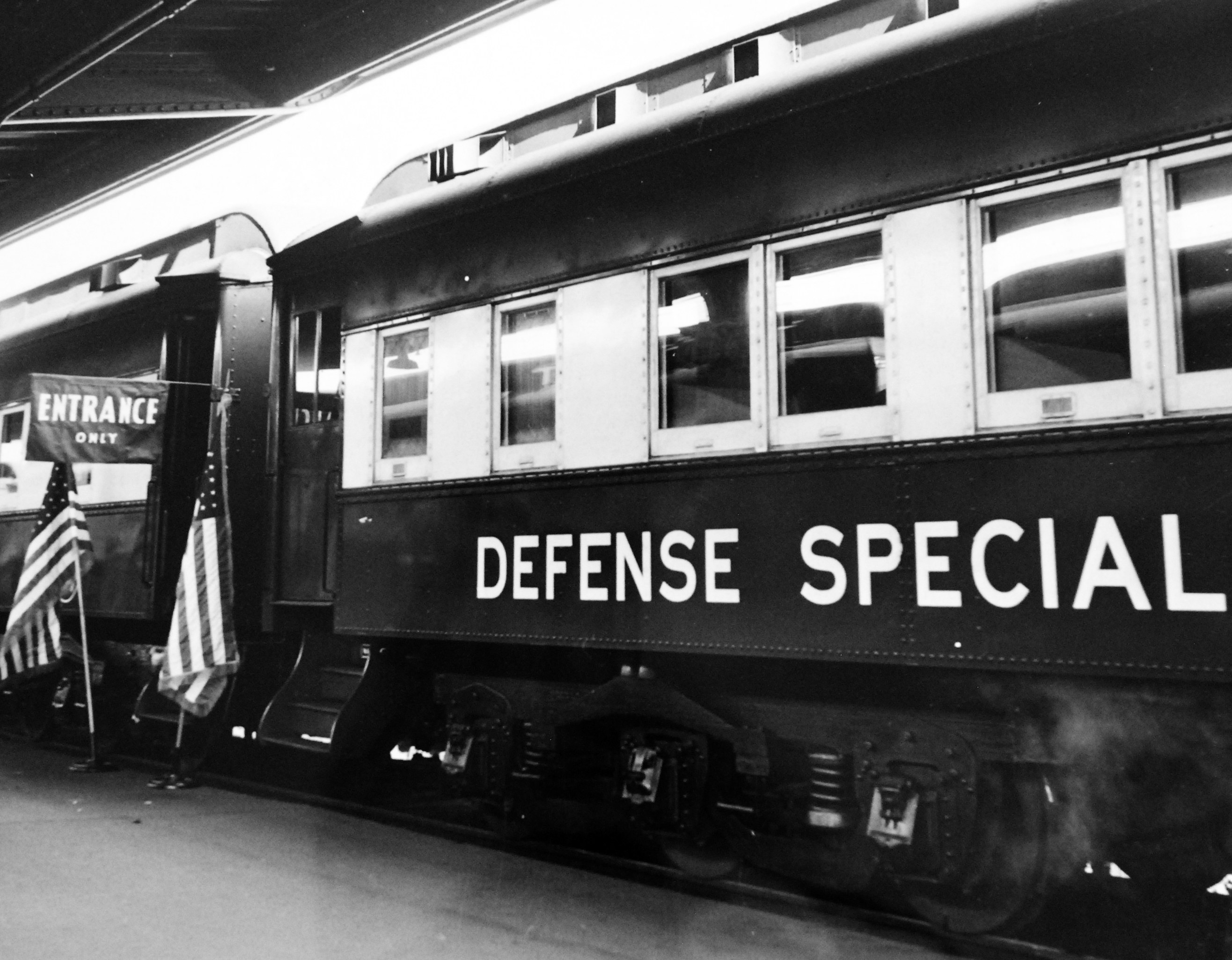 Schwarzes und weißes Foto eines Zuges mit der Aufschrift "Defense Special", der auf den Eisenbahnschienen geparkt ist, mit zwei Fahnen auf der linken Seite und einer Decke oben.