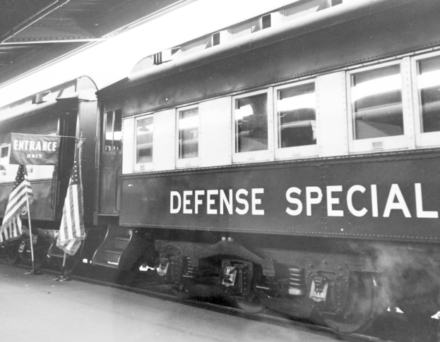 Schwarzes und weißes Foto eines Zuges mit der Aufschrift "Defense Special", der auf den Eisenbahnschienen geparkt ist, mit zwei Fahnen auf der linken Seite und einer Decke oben.