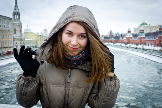 Eine Frau in einer Jacke steht vor einer gefrorenen Wasserfläche, mit Gebäuden und fahrenden Fahrzeugen auf beiden Seiten und einem Himmel im Hintergrund.