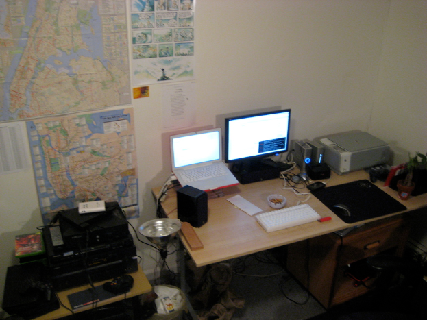 Ein Raum mit einem Tisch, auf dem ein Laptop, ein Lautsprecher, ein Monitor, eine Tastatur, ein Ball, eine Maus, ein Scanner und Papiere liegen; ein weiterer Tisch mit Elektronikgeräten in der unteren linken Ecke; und eine Wand mit Charts und Postern im Hintergrund.