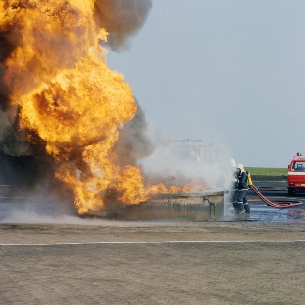 Feuerwehrlöschfahrzeug steht in Flammen an der Stra√e mit zwei Helmet tragenden Personen, die Rohre halten, einem Fahrzeug im Hintergrund und dem Himmel.