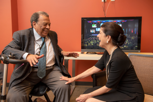Ein Mann und eine Frau sitzen sich in Stühlen gegenüber und unterhalten sich, mit einem Fernseher auf einem Tisch dazwischen, vor einer orangen Wand.