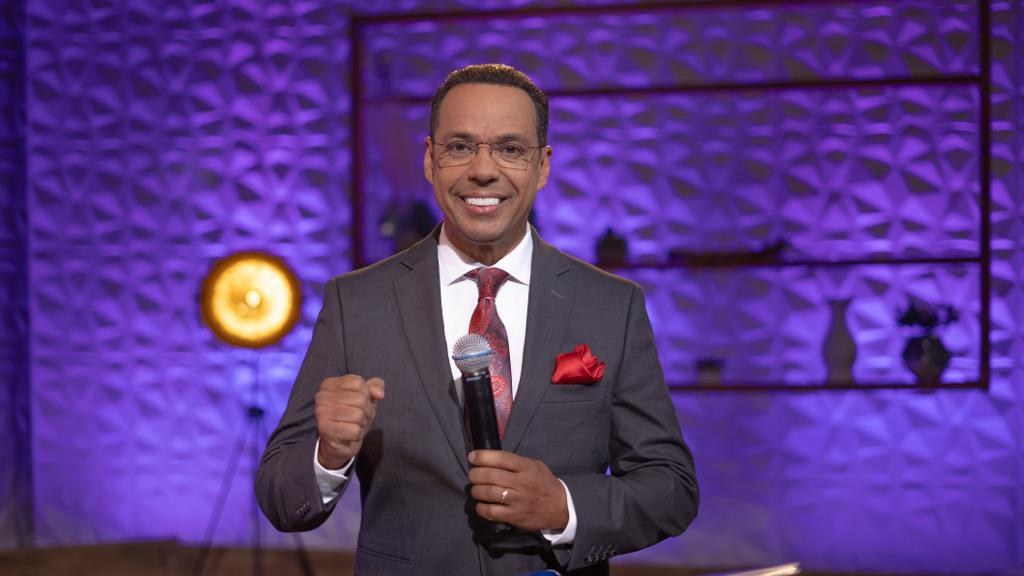 Stephen Colbert, Moderator von 'The Daily Show with Trevor Noah', steht auf einer Bühne und hält ein Mikrofon in der Hand, trägt einen Anzug, eine Krawatte und eine Brille, hinter ihm ist ein Scheinwerfer und ein Ständer zu sehen.