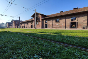 Eine Bahnschiene verläuft durch ein grasbewachsenes Feld neben einem Backsteingebäude mit Fenstern und Türen, umgeben von Strommästen mit Drähten, Bäumen und einigen Menschen, in der Lage Auschwitz-Birkenau-Konzentrationslager in Polen.