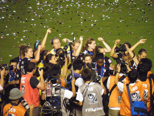 Eine Gruppe von Menschen auf einem Fußballfeld, einige halten Kameras und tragen Taschen, mit verstreuten Papieren im Hintergrund.