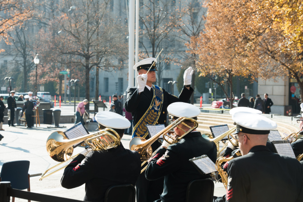 Eine Gruppe uniformierter Männer, die Trompeten spielen, bei einem Umzug, mit Zuschauern in Mützen und Handschuhen, Stühlen und einem Geländer im Vordergrund und Bäumen, Gebäuden, Laternenpfählen, Fahrzeugen und Verkehrskegeln im Hintergrund.