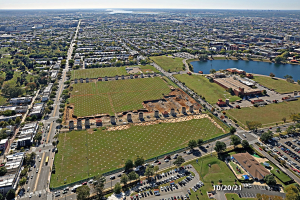 Luftaufnahme einer Stadt mit Gebäuden, Bäumen, Gras, Fahrzeugen, Wasser und Wolken, die einen großen zentralen Platz als Parkplatz zeigt.