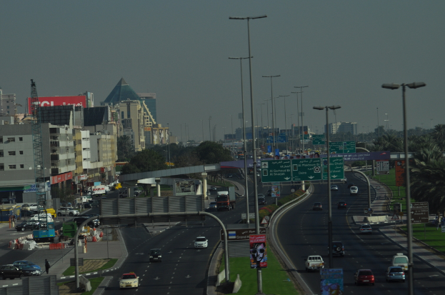 Eine Luftaufnahme einer Stadt mit Gebäuden, Türmen, Straße, Kraftfahrzeugen, Werbetafeln, Schildern, Straßeleuchten, Bäumen, Menschen und Verkehrskegeln.