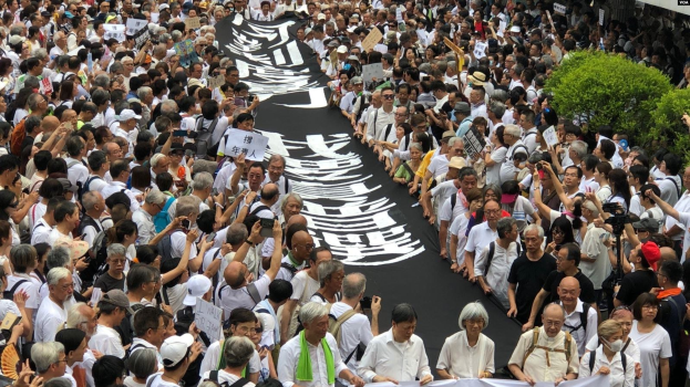 Eine große Gruppe von Menschen marschiert auf der Straße, hält Protestschilder und Banner in der Hand, mit Topfpflanzen auf der rechten Seite und einem Gebäude im Hintergrund.