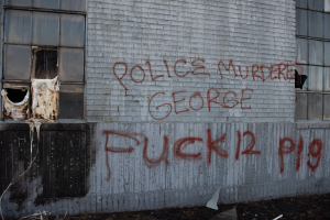 Graffiti an einer Hauswand mit der Aufschrift "Polizeimord George Fuck 12 P19" mit Fenstern und verstreuten Gegenständen davor.