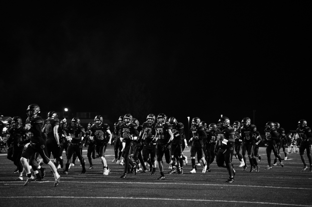 Eine Gruppe von Fußballspielern in Helmen, die nachts auf das Feld laufen, umgeben von Pfosten und Lichtern, in Schwarz-Weiß.