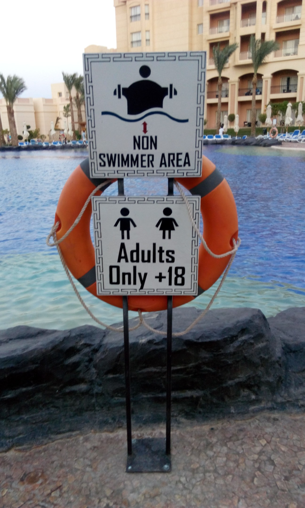 Ein Schwimmbad mit einem "Non Swimmer Area Adults Only"-Schild im Vordergrund, umgeben von Bäumen, Gebäuden und einem klaren blauen Himmel, mit einem Rettungsring in der Mitte des Pools.