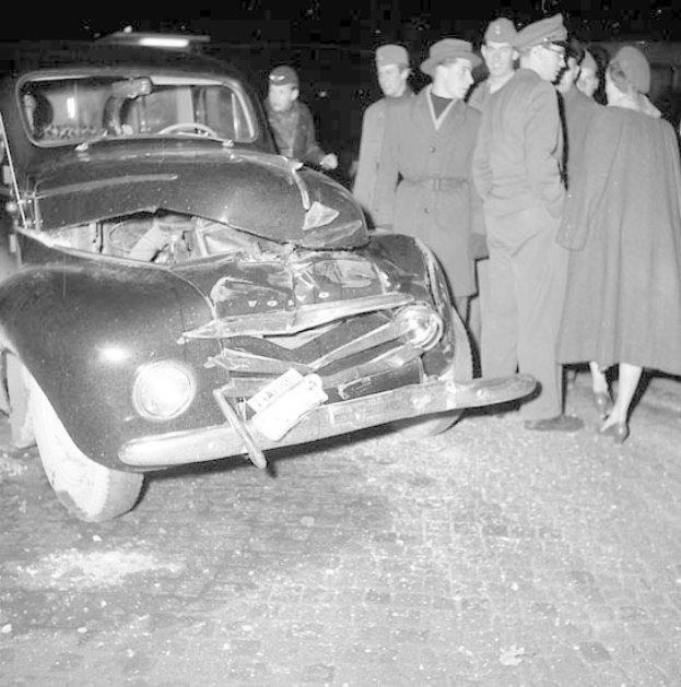 Schwarz-weißes Foto eines stark beschädigten Autos am Straßenrand mit eingedellter Motorhaube und zersplitterten Fenstern, umgeben von Menschen, die es inspizieren.