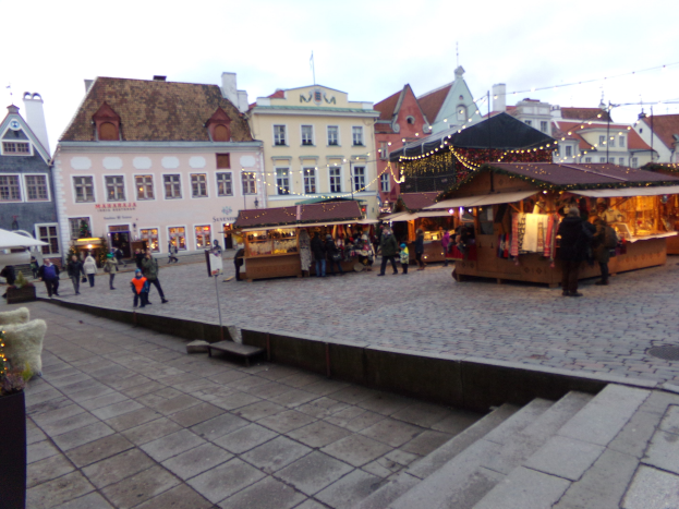Ein geschäftiger Weihnachtsmarkt in Tallinn, Estland mit Menschen um dekorierte Buden, festliche Lichter und Topfpflanzen, vor Wolkenhimmel und Gebäuden mit Fenstern.