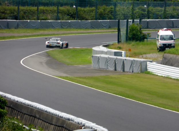 Ein Rennwagen fährt eine Rennstrecke entlang, im Hintergrund ist ein Polizeiwagen zu sehen, umgeben von Grünflächen und Absperrungen, mit Text am unteren Bildrand.