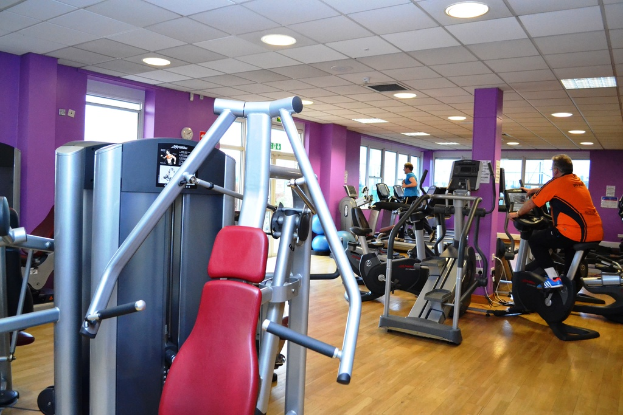 Ein Fitnessstudio mit mehreren Personen, die auf verschiedenen Geräten trainieren, mit einer Decke und Lampen oben im Bild.