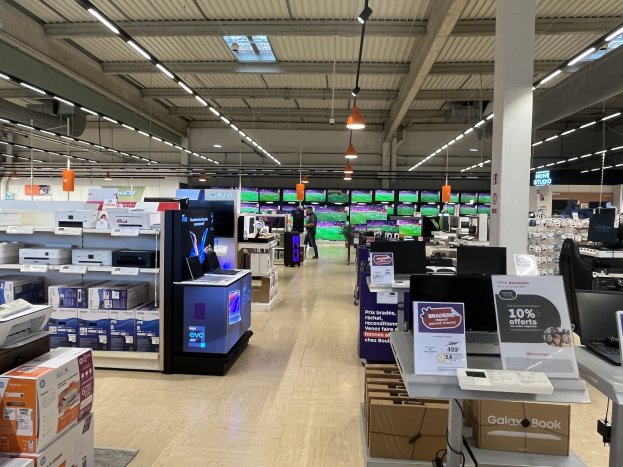 Ein großer Raum in einem Walmart-Laden, der mit Elektronik gefüllt ist, die in Regalen angeordnet ist, einschließlich Computer, Drucker und Kartons, mit Menschen auf dem Boden, einer Säule, Schildern, Deckenlampen und Deckenventilatoren.