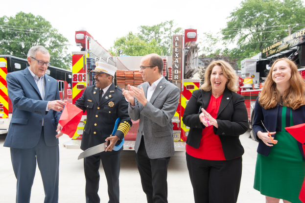 Eine Gruppe von Menschen steht vor einem Feuerwehrauto, klatscht und lacht, mit zwei Personen, die eine Schere und ein rotes Tuch halten, bei einer Eröffnungszeremonie der Feuerwehr Alexandria, mit Bäumen und einem klaren blauen Himmel im Hintergrund.