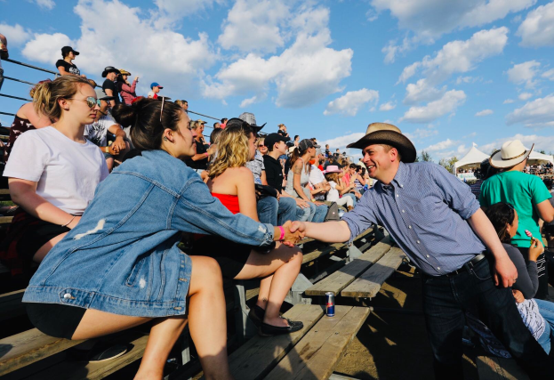 Ein Mann in einem blauen Hemd und schwarzen Hosen gibt einer Frau in unbekannter Kleidung die Hand, vor einem Publikum auf Bänken sitzend und weißen Zelten, Bäumen und einem blauen und weißen Himmel im Hintergrund.