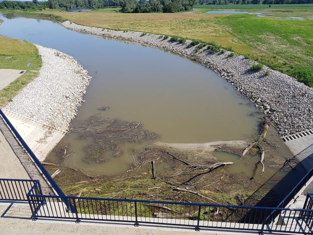Ein Fluss fließt durch ein grünes Feld neben einer Brücke mit Geländern, Steinen, Gras und Bäumen im Hintergrund unter einem bewölkten Himmel, Teil des Mississippi-River-Restoration-Projekts.