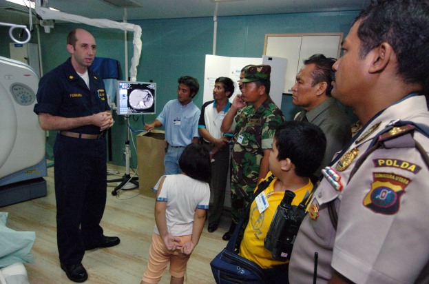 Eine Gruppe von Menschen in einem Krankenhauszimmer mit einem Mann, der ein Mikrofon hält, einem drapierten Bett, medizinischen Geräten, einem Röntgengerät, einer Texttafel, einer Tür und Deckenleuchten.