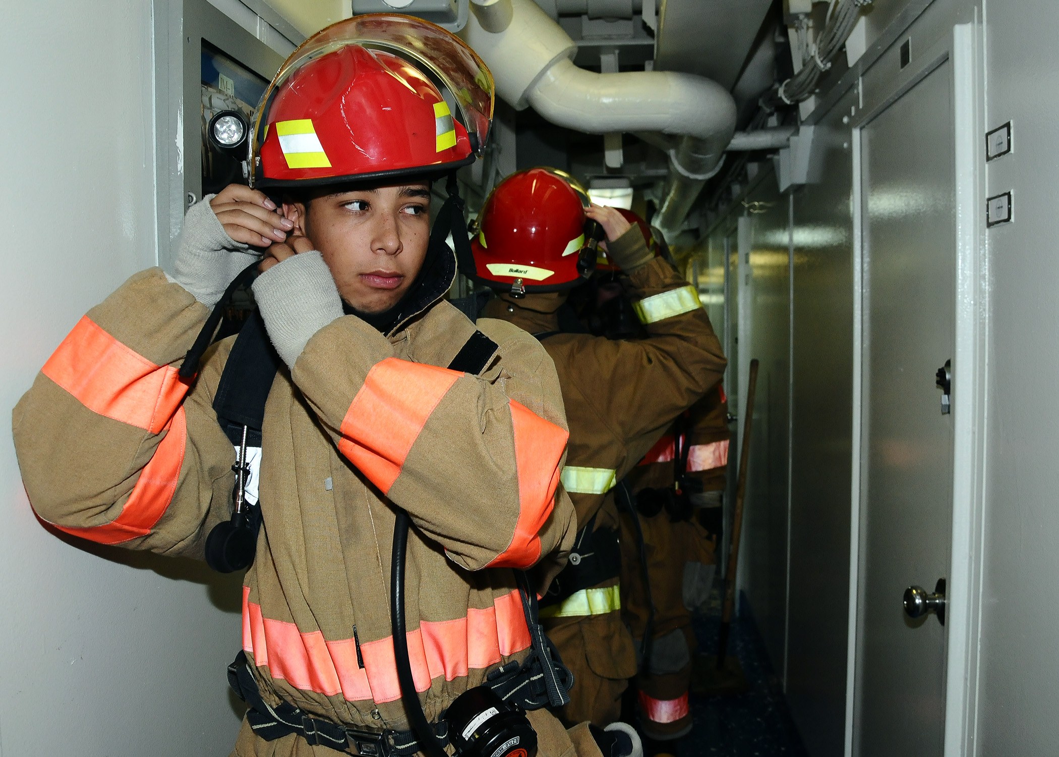Eine Gruppe von Feuerwehrleuten in Uniform, die in einem Raum mit einer Tür auf der rechten Seite und einer Wand auf der linken Seite stehen, während einer Übung mit Rohren und Equipment im Hintergrund.