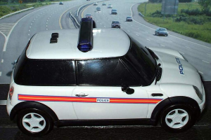 Ein weißer Polizeiwagen steht im Vordergrund, während sich im Hintergrund mehrere Autos auf der Straße bewegen, umgeben von Gras und Bäumen auf beiden Seiten.